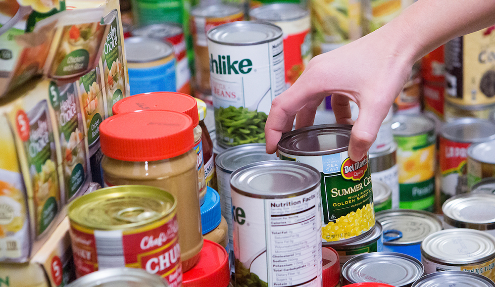 A hand reaches to grab a can of corn on a pile of shelf-stable and canned food at a food pantry. A hand reaches to grab a can of corn on a pile of shelf-stable and canned food at a food pantry.