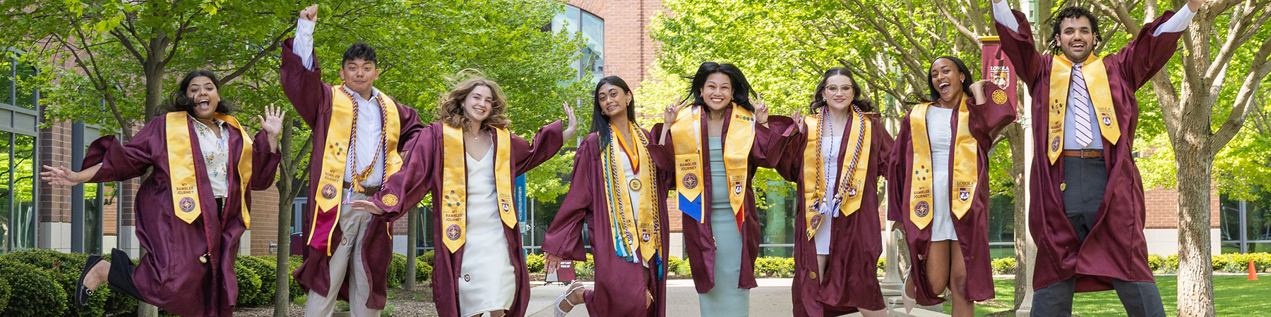 A joyful group of diverse Loyola graduates wearing maroon caps and gowns leap into the air on a sunny day on the Lake Shore campus.