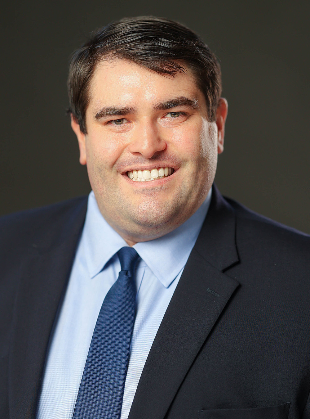 Portrait of a smiling man wearing a dark suit and blue tie against a dark background Portrait of a smiling man wearing a dark suit and blue tie against a dark background