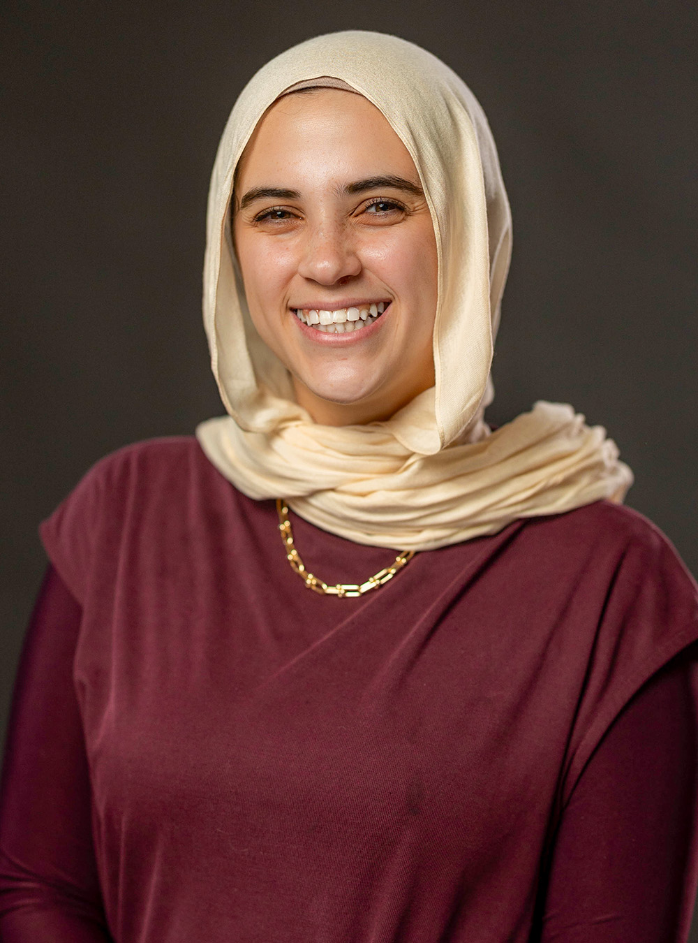 Portrait of a smiling woman wearing a light-colored headscarf and maroon top against a dark background Portrait of a smiling woman wearing a light-colored headscarf and maroon top against a dark background