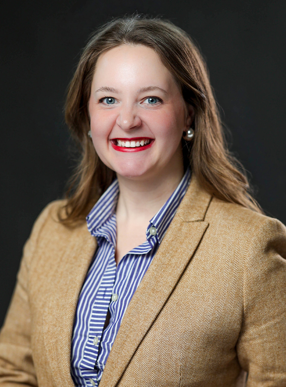 Portrait of a smiling woman with long light brown hair wearing a tan blazer and striped shirt against a dark background Portrait of a smiling woman with long light brown hair wearing a tan blazer and striped shirt against a dark background