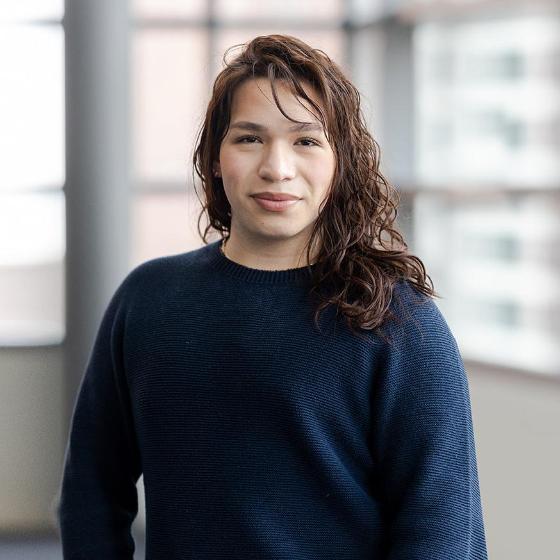 Arrupe College student standing indoors in front of large windows, wearing a navy sweater and looking confidently at the camera.