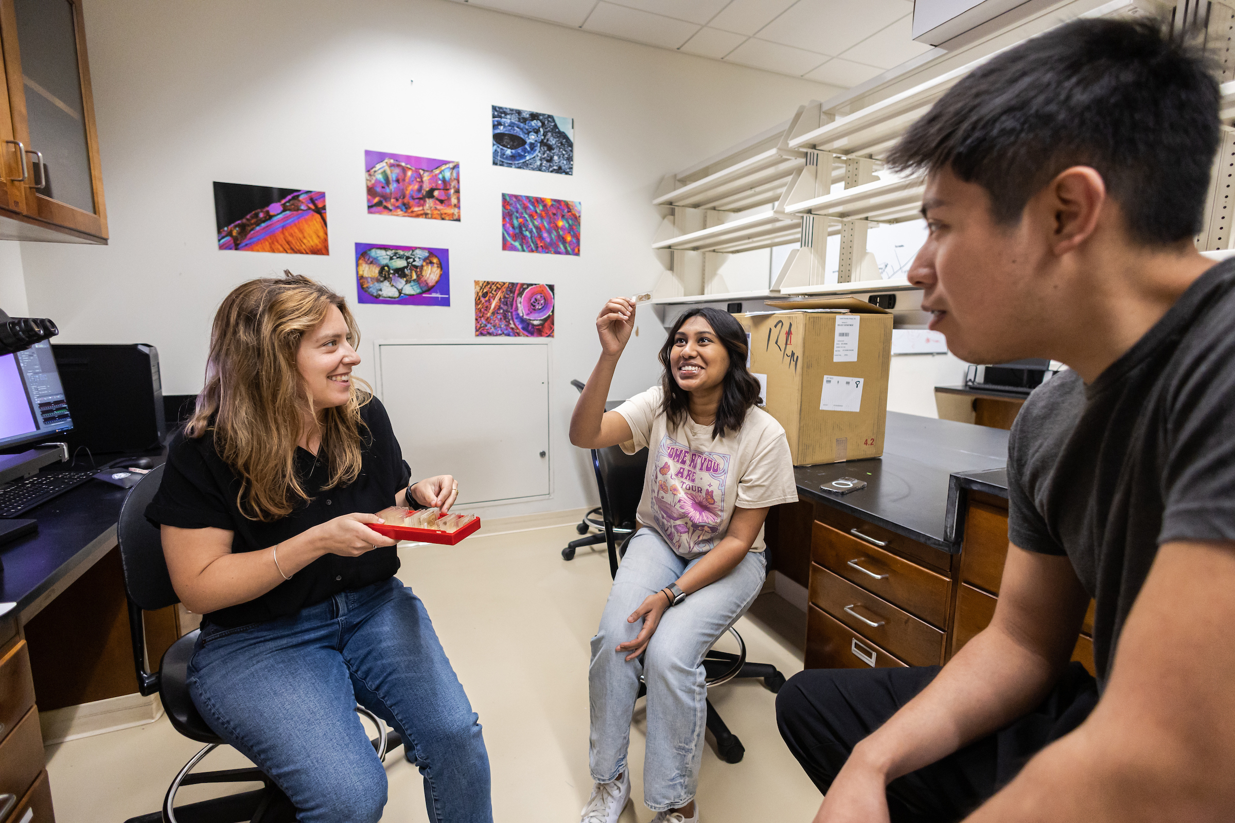 Megan Whitney speaks to Pranati and Eduardo as Pranati holds up a microscope slide and looks at the sample against the light