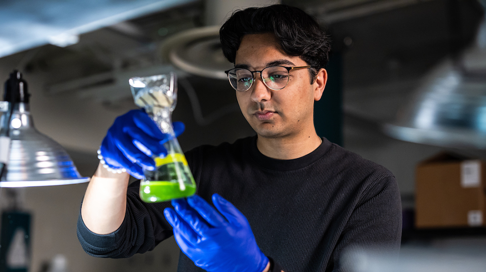 Arivu Kapoor holds a glass beaker containing a green liquid in a chemistry lab
