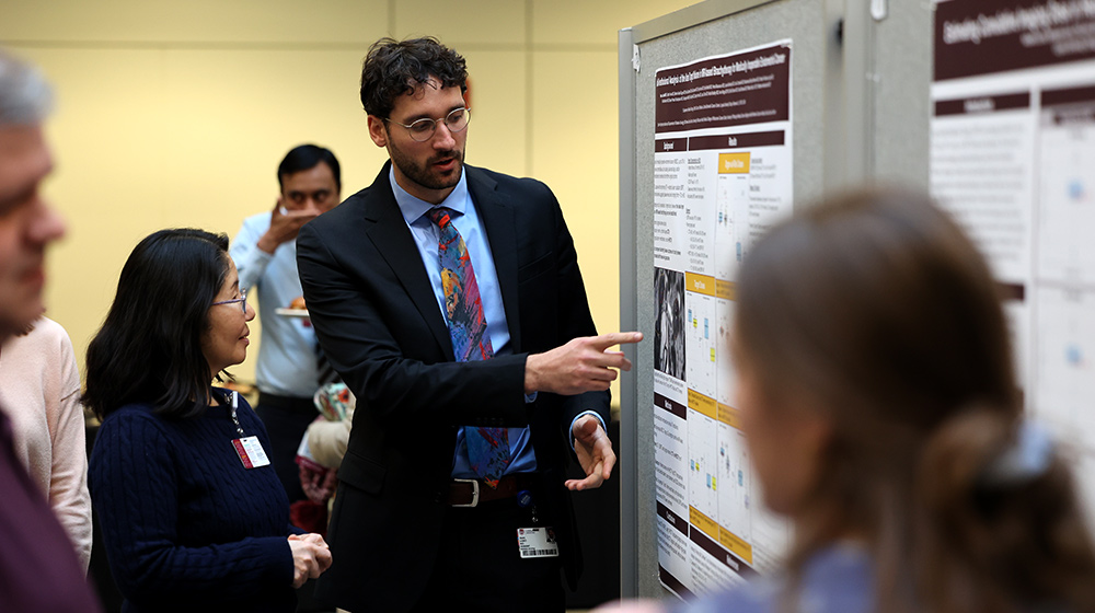 A presenter in professional attire points to information on a research poster while attendees stand nearby reviewing additional displayed academic materials. A presenter in professional attire points to information on a research poster while attendees stand nearby reviewing additional displayed academic materials.