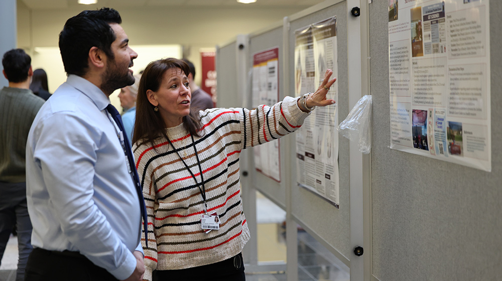 Two individuals stand closely together as one gestures toward a research poster on a display board during an indoor academic event. Two individuals stand closely together as one gestures toward a research poster on a display board during an indoor academic event.