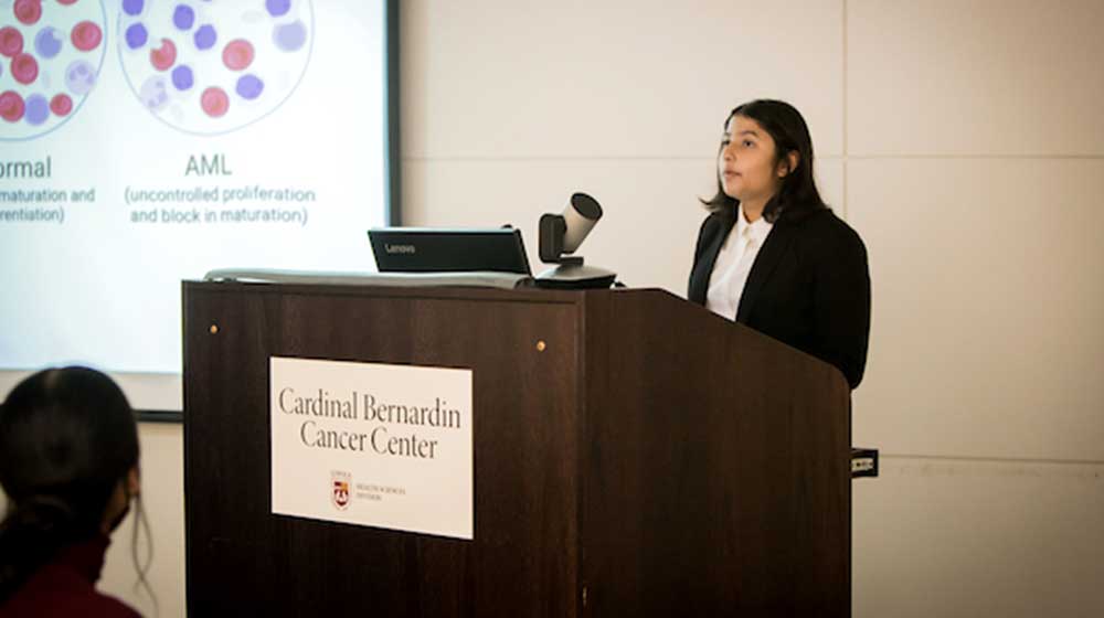 Presenter stands at a podium delivering a scientific lecture with slides displayed behind them at the Cardinal Bernardin Cancer Center.