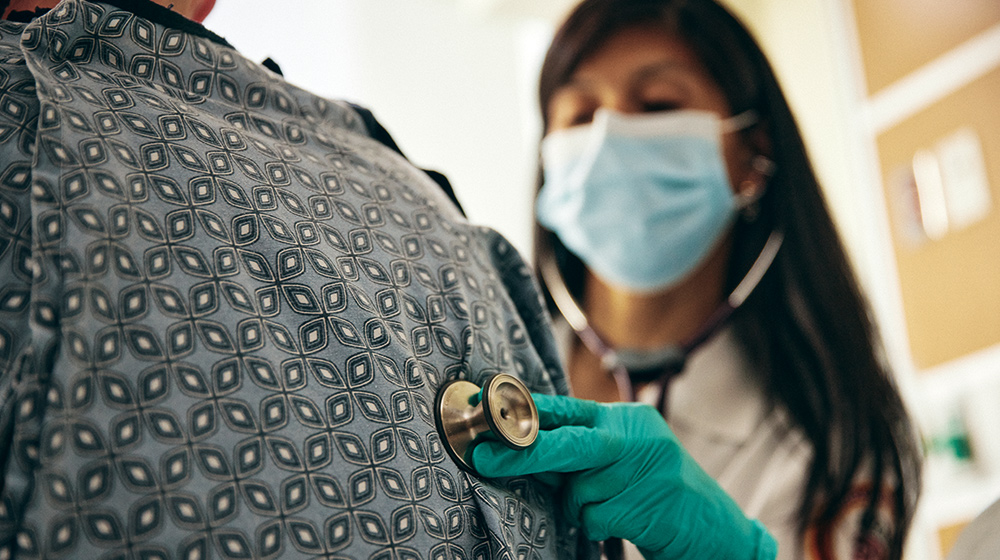 Gloved medical professional places a stethoscope on a patterned gown during a clinical exam, with examination room equipment visible in the background.