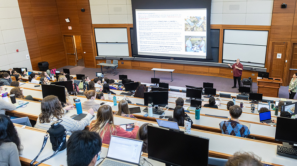 Students sit in a tiered lecture hall while an instructor presents slides on a large screen during a university class. Students sit in a tiered lecture hall while an instructor presents slides on a large screen during a university class.