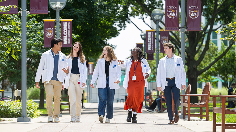 A group of students wearing white lab coats walk together along a tree-lined campus pathway with Loyola welcome banners. A group of students wearing white lab coats walk together along a tree-lined campus pathway with Loyola welcome banners.