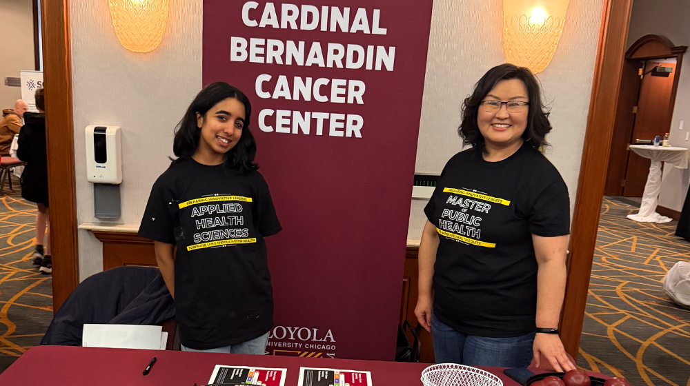 Two individuals stand at an information table with brochures in front of a Cardinal Bernardin Cancer Center display wearing program‑themed shirts. Two individuals stand at an information table with brochures in front of a Cardinal Bernardin Cancer Center display wearing program‑themed shirts.