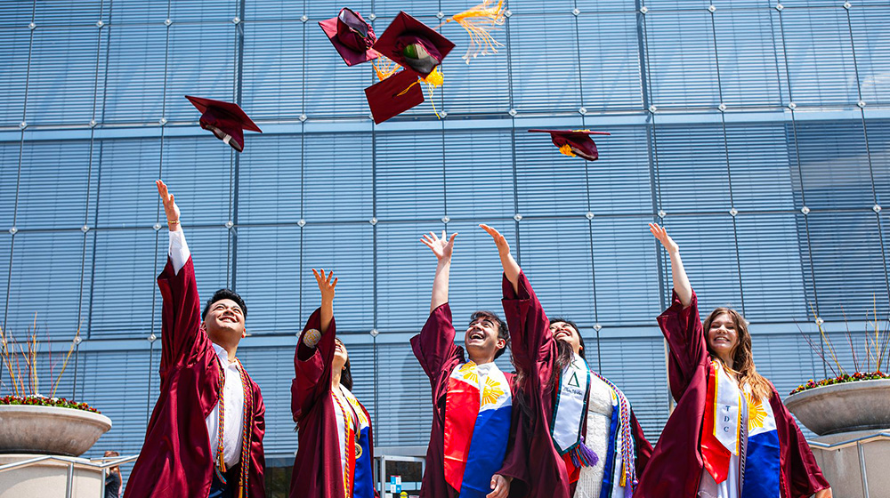 A group of 2024 graduates throw their caps in the air in celebration standing in front of the Information Commons building