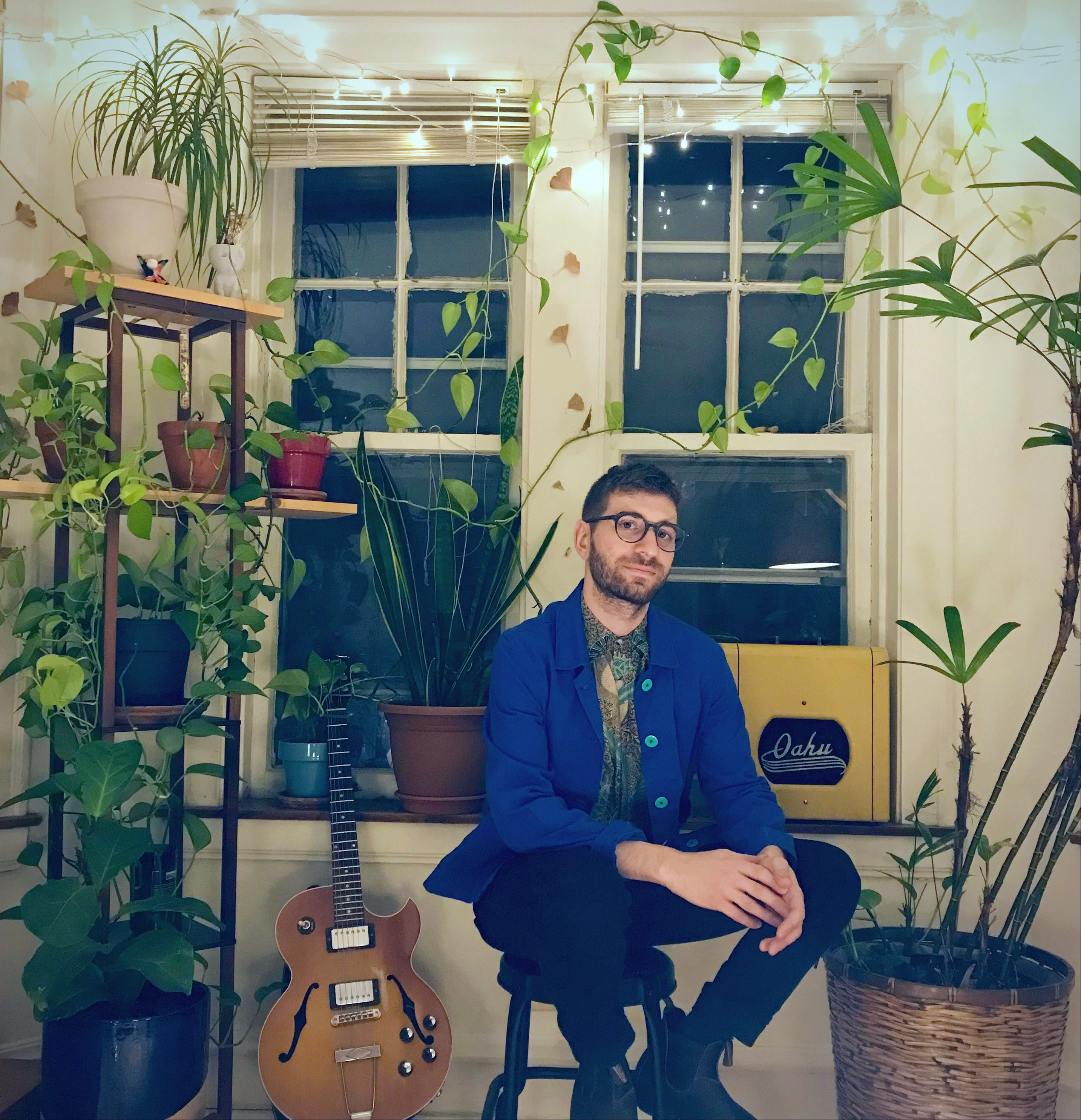 Matt Gold sitting on a stool in front of a window, surrounded by tall green plants