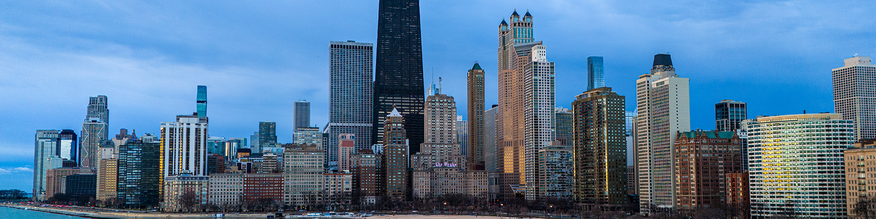 Panoramic view of Chicago skyline along Lake Michigan, showing high-rise buildings and waterfront with city lights under a cloudy sky.