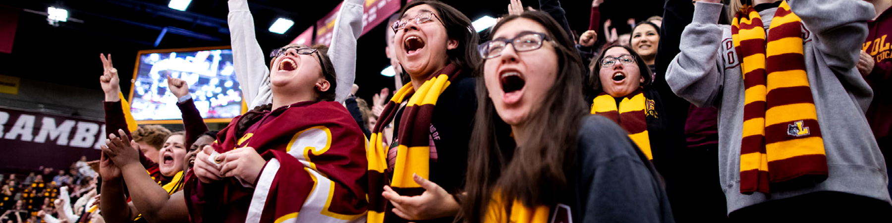 Students in maroon and gold apparel cheer in a packed arena, wearing scarves and jerseys during a collegiate sporting event.