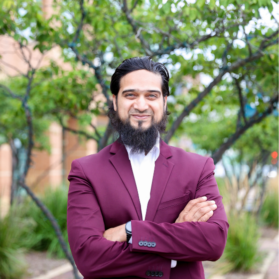 A smiling man with dark brown hair pulled back and with a full beard, wearing a maroon blazer over a white top, arms folded, standing in front of a school campus. A smiling man with dark brown hair pulled back and with a full beard, wearing a maroon blazer over a white top, arms folded, standing in front of a school campus.