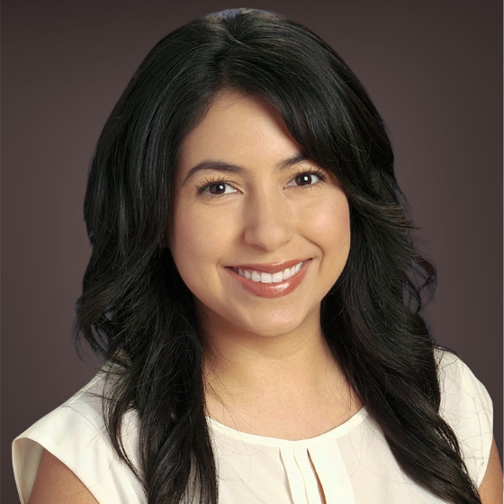 A smiling woman with wavy brown hair, wearing a white blouse, against a solid dark grey background. A smiling woman with wavy brown hair, wearing a white blouse, against a solid dark grey background.