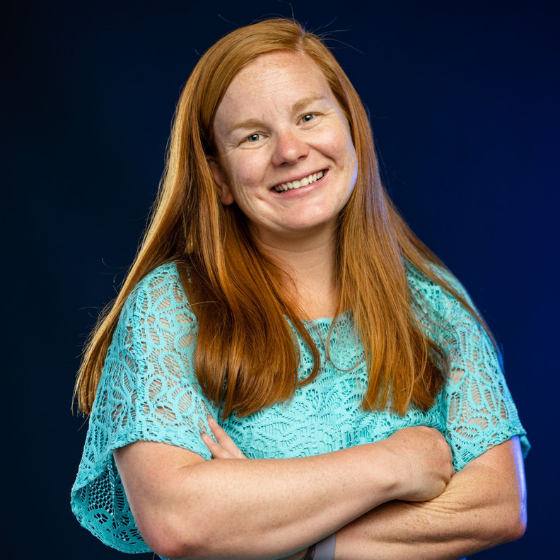 Woman with long, reddish hair smiling and wearing a teal short-sleeved top, against a solid black background. Woman with long, reddish hair smiling and wearing a teal short-sleeved top, against a solid black background.