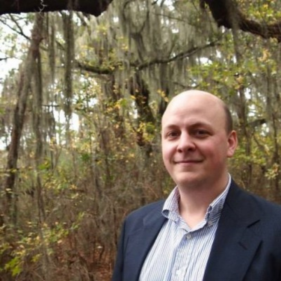 A bald man smiling slightly wearing a striped button up shirt underneath an open navy blue blazer, in front of a forest.