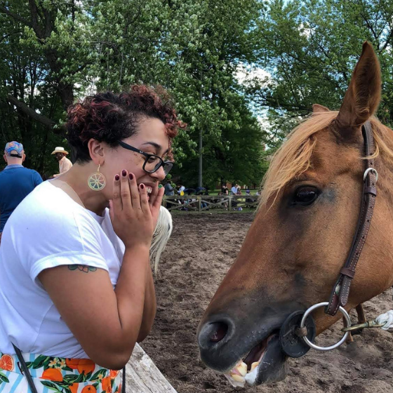 A woman with short, curly hair smiling at a brown horse, who is also smiling. The woman is wearing glasses, a white top, and a patterned skirt. A woman with short, curly hair smiling at a brown horse, who is also smiling. The woman is wearing glasses, a white top, and a patterned skirt.