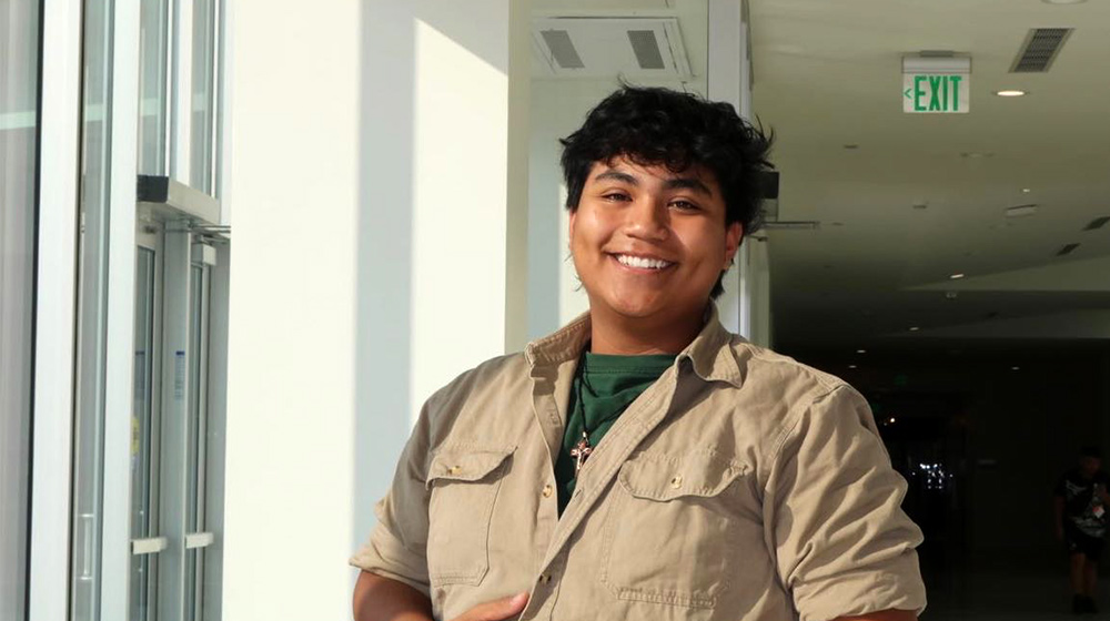 Person stands in a sunlit indoor hallway near glass doors, wearing a button-up shirt, facing the camera with exit sign.