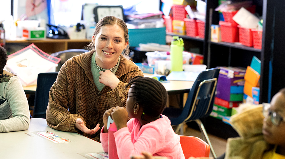 Adult educator sits at classroom table, engaging young students in discussion, with learning materials and colorful bins visible behind them. Adult educator sits at classroom table, engaging young students in discussion, with learning materials and colorful bins visible behind them.
