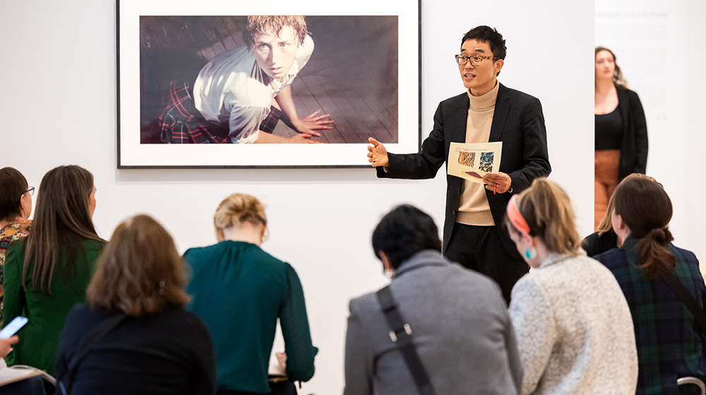 Presenter stands before seated audience, gesturing while holding paper during talk beneath large framed photograph in gallery exhibition space.