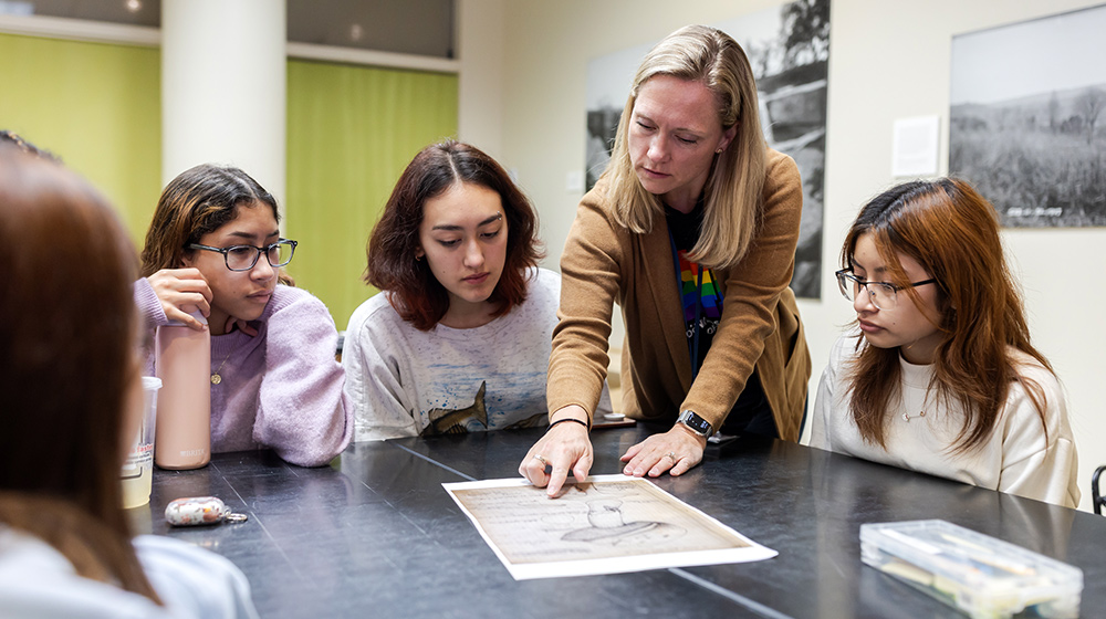 Educator leans over table pointing to historical print while four students closely examine materials during small-group learning session.