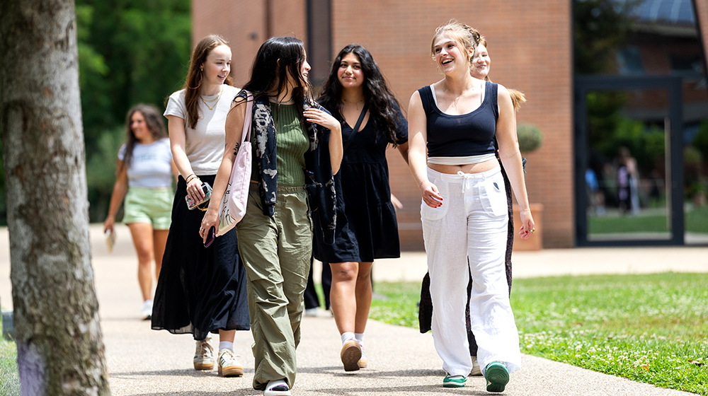 Group of students walk together along campus sidewalk between grass and brick buildings on sunny day carrying bags casually.