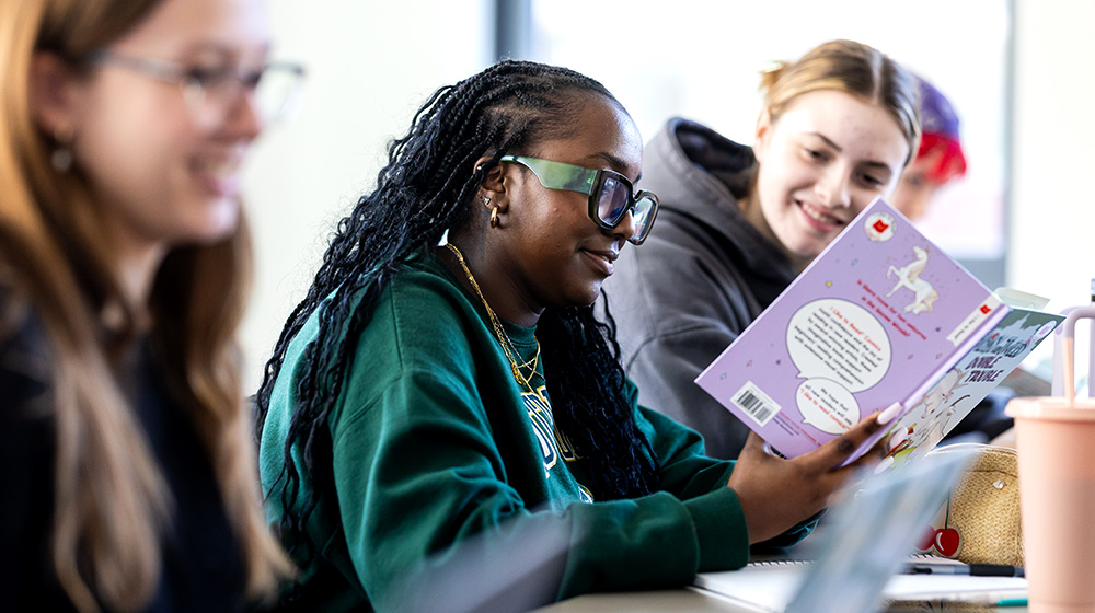 Students sit at a table reading a colorful book during a collaborative study session with notebooks, laptops, and drinks. Students sit at a table reading a colorful book during a collaborative study session with notebooks, laptops, and drinks.