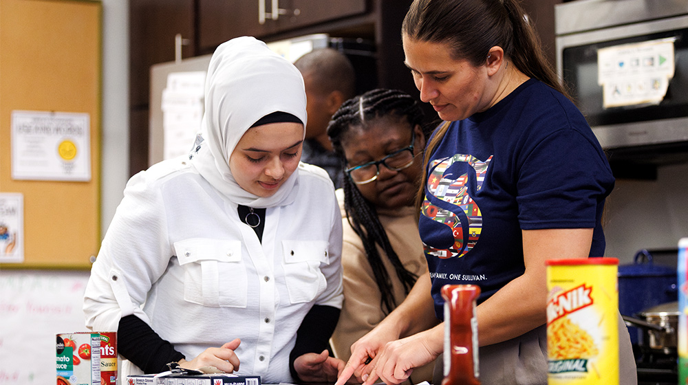 Participants prepare food together in a classroom kitchen, following instructions, with canned ingredients, condiments, appliances visible nearby countertops. Participants prepare food together in a classroom kitchen, following instructions, with canned ingredients, condiments, appliances visible nearby countertops.