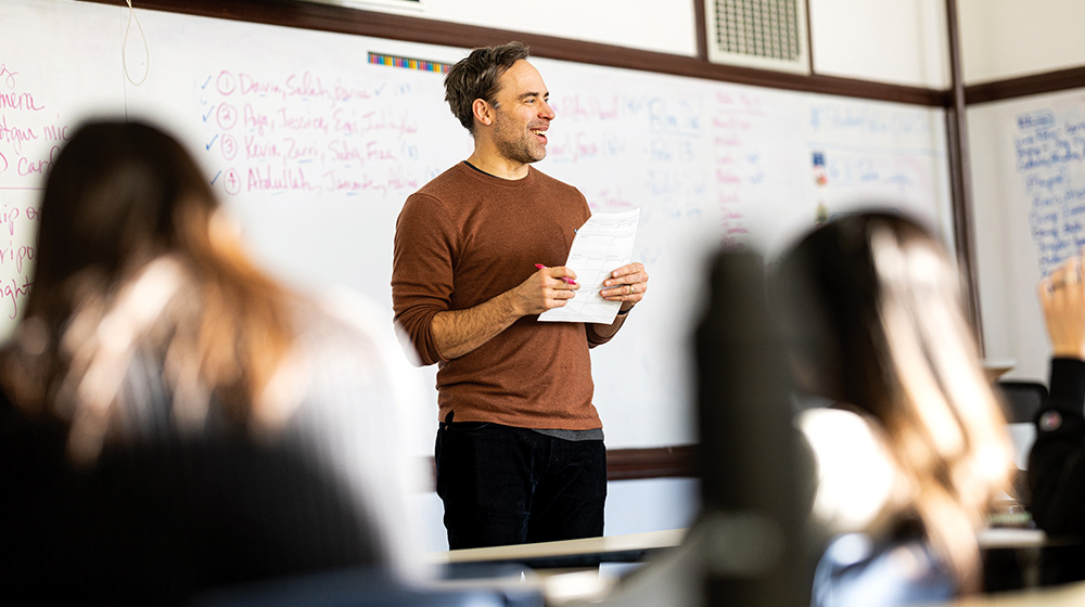 Instructor stands at front of classroom holding papers and marker, speaking to seated learners with whiteboard notes behind visible. Instructor stands at front of classroom holding papers and marker, speaking to seated learners with whiteboard notes behind visible.