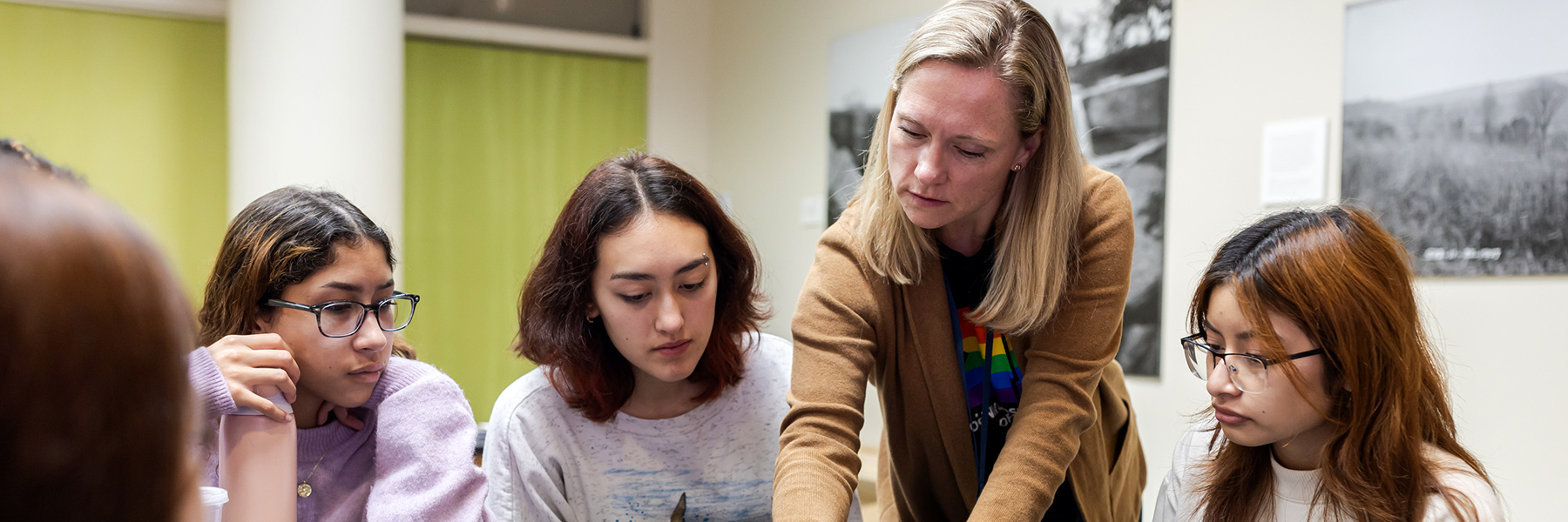 Adult educator guiding four students as they collaborate at a table in a classroom with photos on walls displayed.