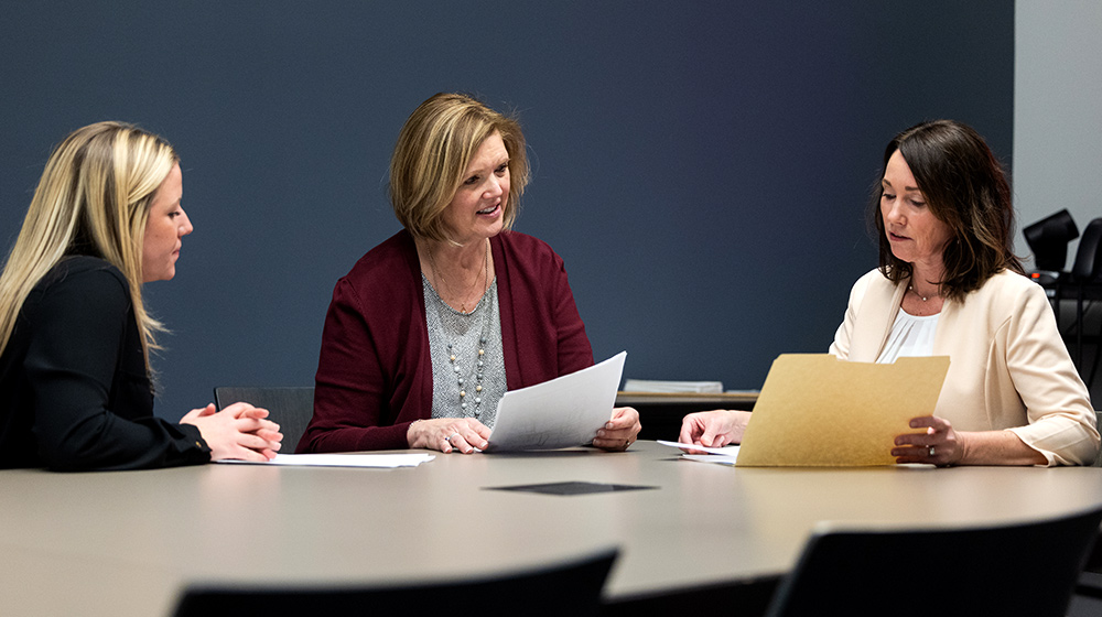 Three people review printed documents together at a conference table in a modern office meeting room with blue wall backdrop.