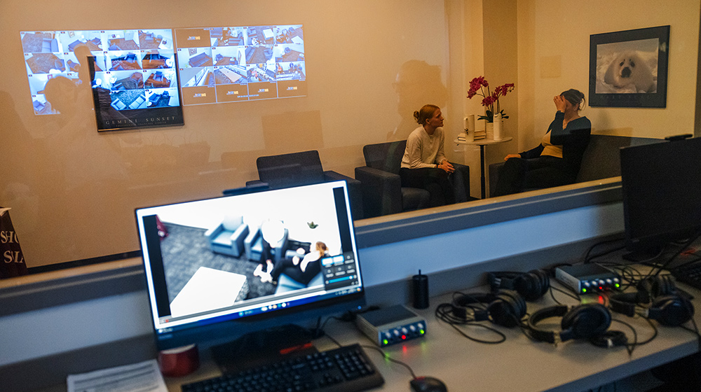 Observation room shows simulation lab with control desk, monitors, headsets, and two participants seated behind glass reviewing projected scenarios.