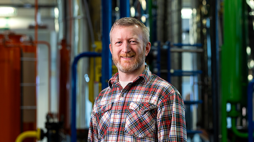Person wearing a plaid shirt stands in an industrial facility surrounded by metal pipes, machinery, and large processing equipment.
