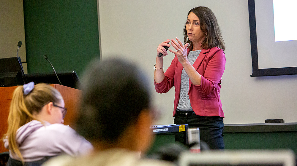 A woman presents a lecture, gesturing with a remote while students listen in 