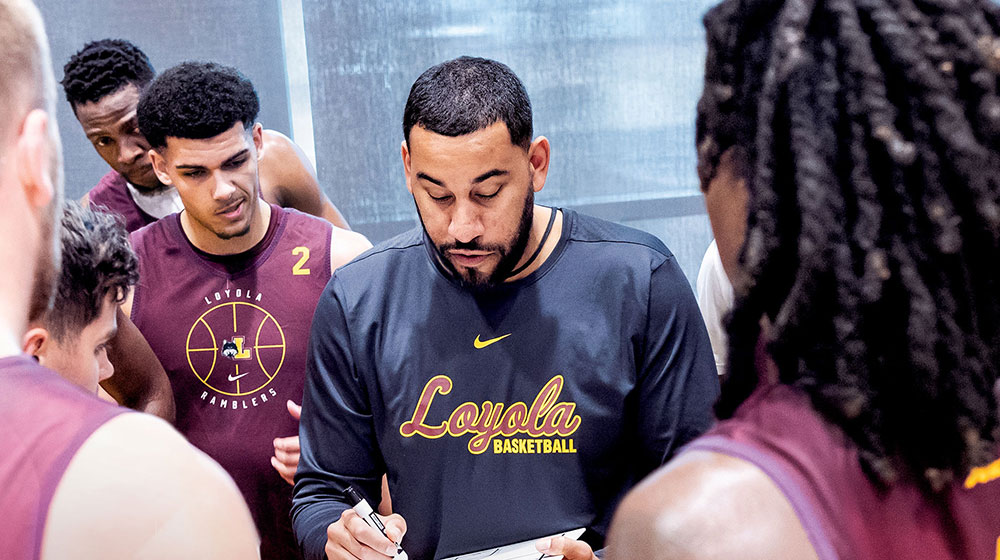 Loyola men's basketball coach Drew Valentine in a huddle with his players in their uniforms while he draws plays on a handheld whiteboard.