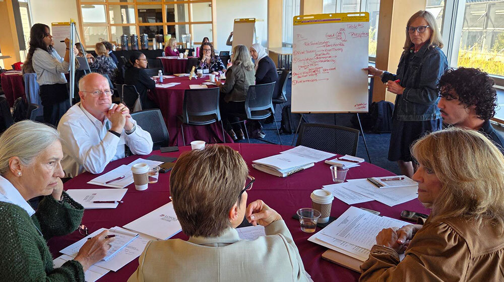 Adults sit at several tables filling a large room and talk. Some stand in front of large white notepads and write. Adults sit at several tables filling a large room and talk. Some stand in front of large white notepads and write.