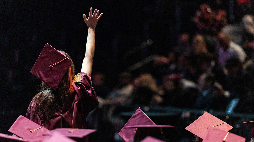 A woman in a maroon robe stands, back to the camera, and raises her right hand during a Commencement ceremony. The heads of other graduates, wearing graduation caps, are visible. A woman in a maroon robe stands, back to the camera, and raises her right hand during a Commencement ceremony. The heads of other graduates, wearing graduation caps, are visible.