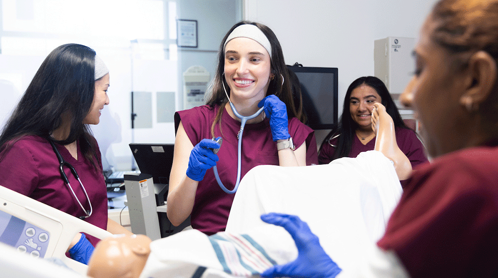 A student in maroon scrubs holds a stethoscope and smiles while standing over a manikin. A student in maroon scrubs holds a stethoscope and smiles while standing over a manikin.