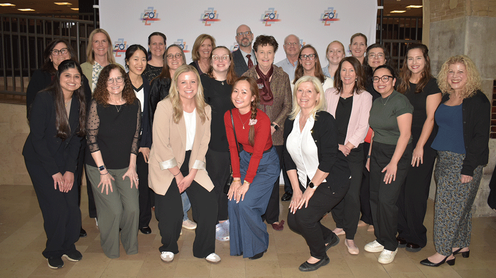 Approximately two dozen people stand together in front of a banner and smile Approximately two dozen people stand together in front of a banner and smile