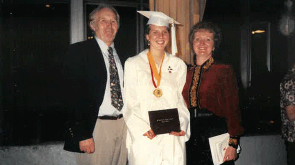 Melissa Murphey stands in white graduation gown with her father standing to her right and her mother to her left. Melissa Murphey stands in white graduation gown with her father standing to her right and her mother to her left.