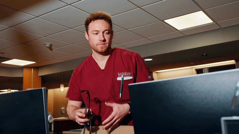 A male nurse in maroon scrubs holds a stethoscope and looks at a computer monitor in an office A male nurse in maroon scrubs holds a stethoscope and looks at a computer monitor in an office