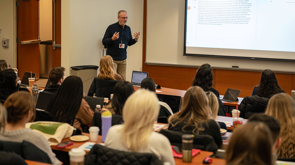 A man stands at the front of a crowded lecture hall and talks to DNP students
