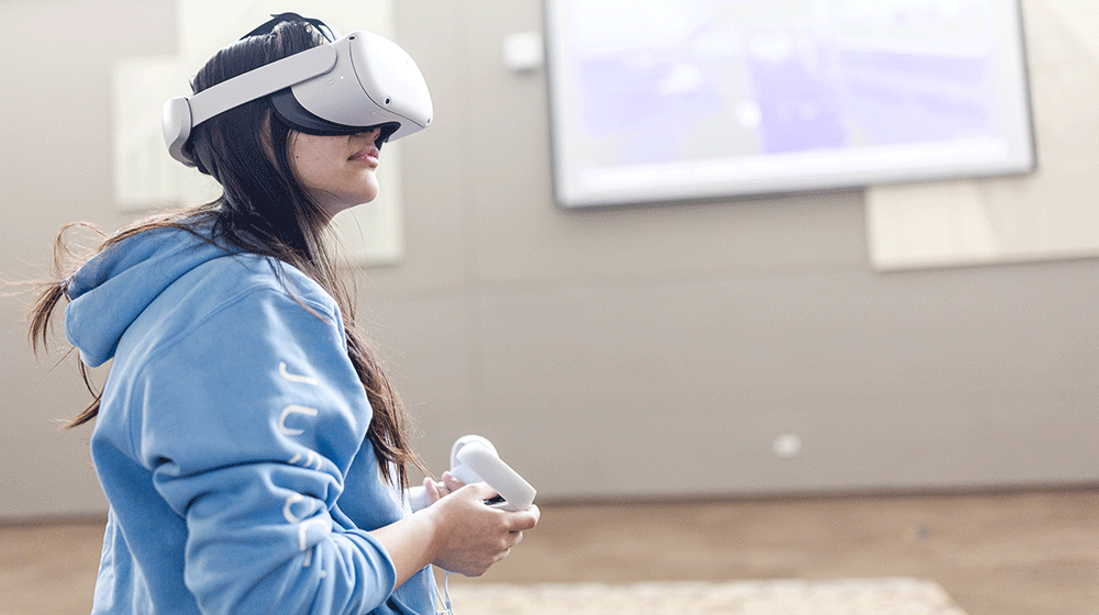 A female student in a blue sweatshirt uses VR equipment