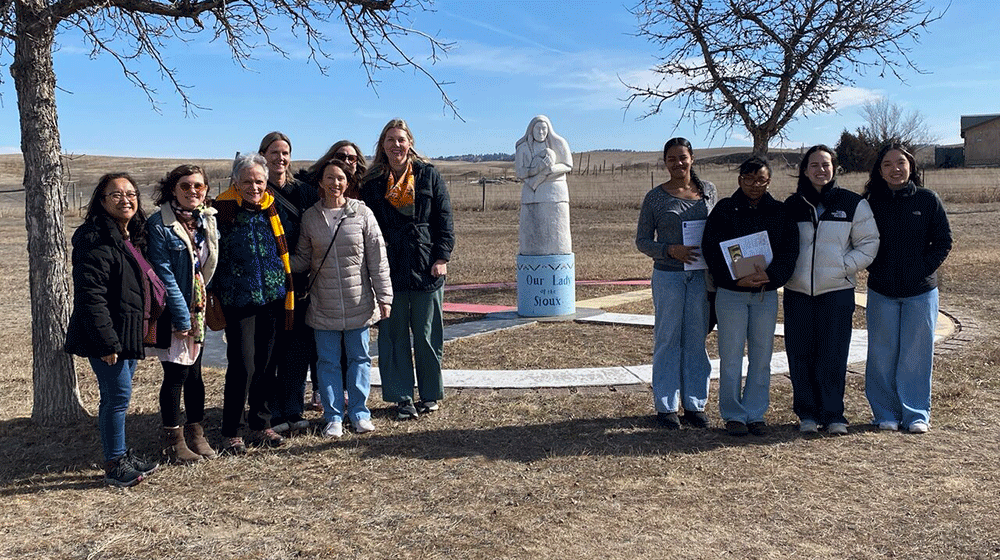 Students and faculty stand on grass in front of a person-sized statue of Mary holding Baby Jesus Students and faculty stand on grass in front of a person-sized statue of Mary holding Baby Jesus