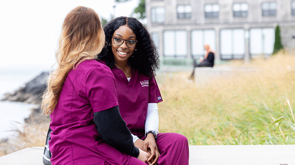 Two students in maroon scrubs sit and talk on the Lake Shore Campus beside Lake Michigan