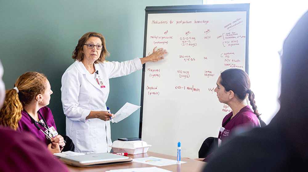 A faculty member in a white nursing coat stands in front of a white board teaching undergraduates in maroon scrubs