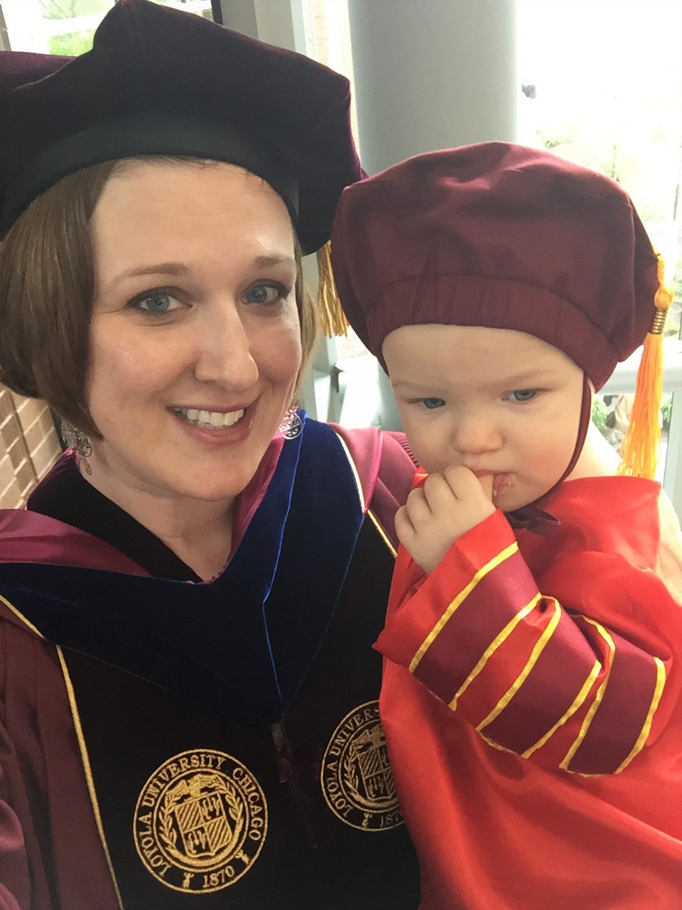 A photo of PhD graduate Merrit Rehn-DeBraal on graduation day, holding her daughter Mila, who is also wearing a PhD graduation cap.